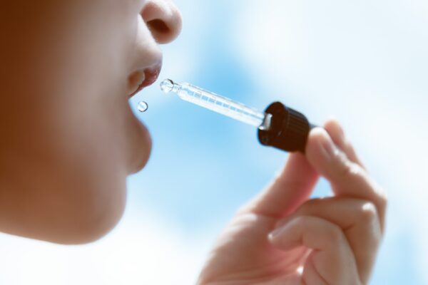 Side profile of a woman tilting her head back as a single drop falls from a dropper toward her parted lips